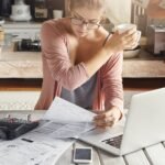 concentrated woman dressed casually calculating bills, sitting at kitchen table with laptop, calculator, papers and mobile, holding white cup and passing it to her husband, who is standing behind her