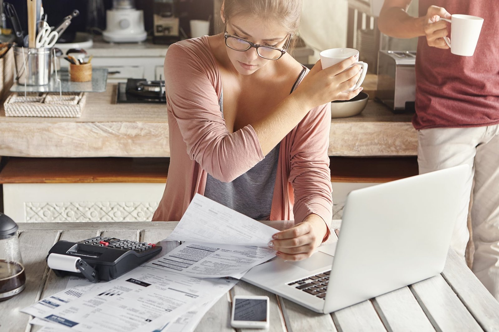 concentrated woman dressed casually calculating bills, sitting at kitchen table with laptop, calculator, papers and mobile, holding white cup and passing it to her husband, who is standing behind her